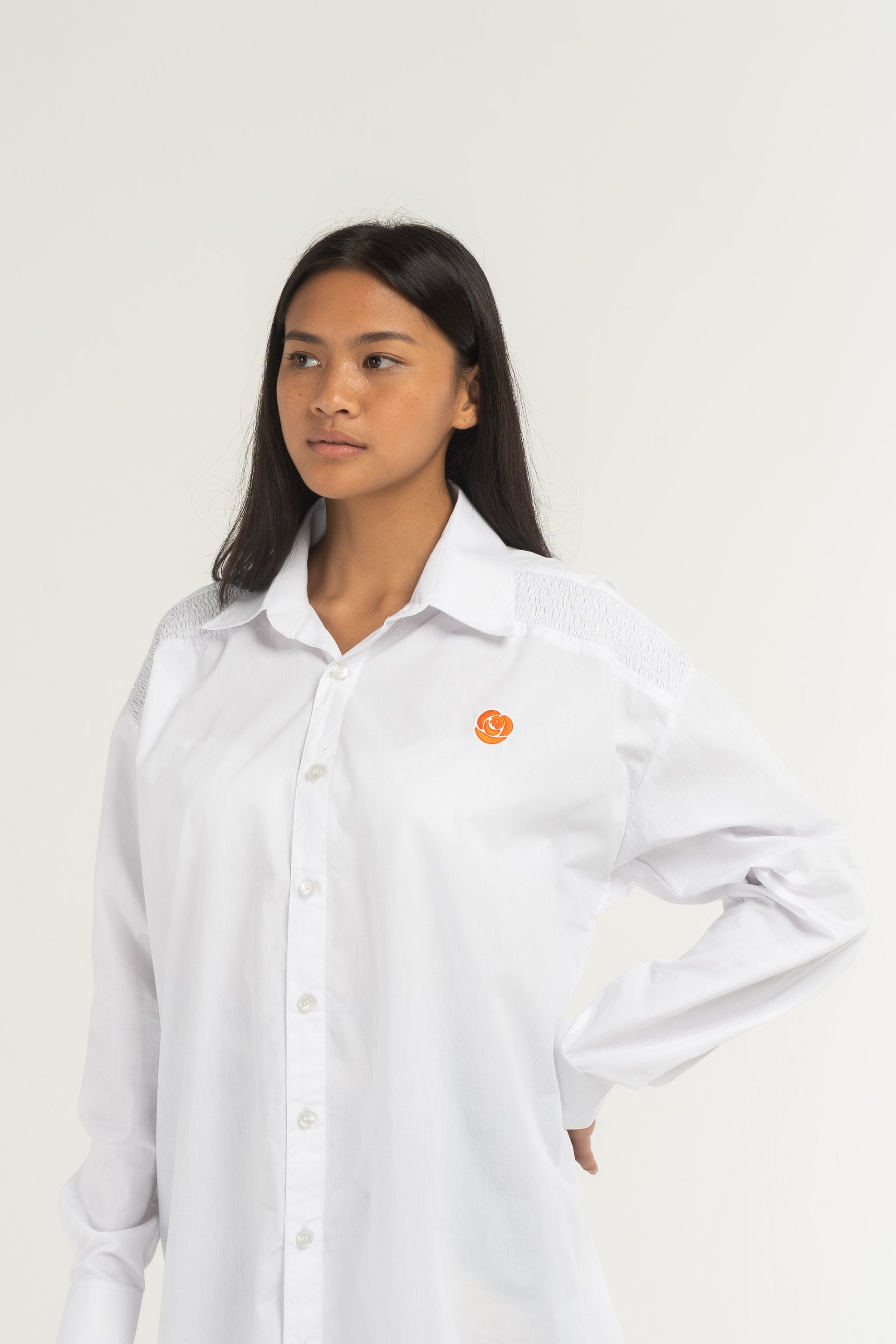 A white Toggle Shirt Dress forms a long silhouette with a curved hem, featuring the Sweet Rose logo hand-embroidered on the front, long-sleeves with a button-up front, a toggle fastening at the back, a structured collar and cuffs.