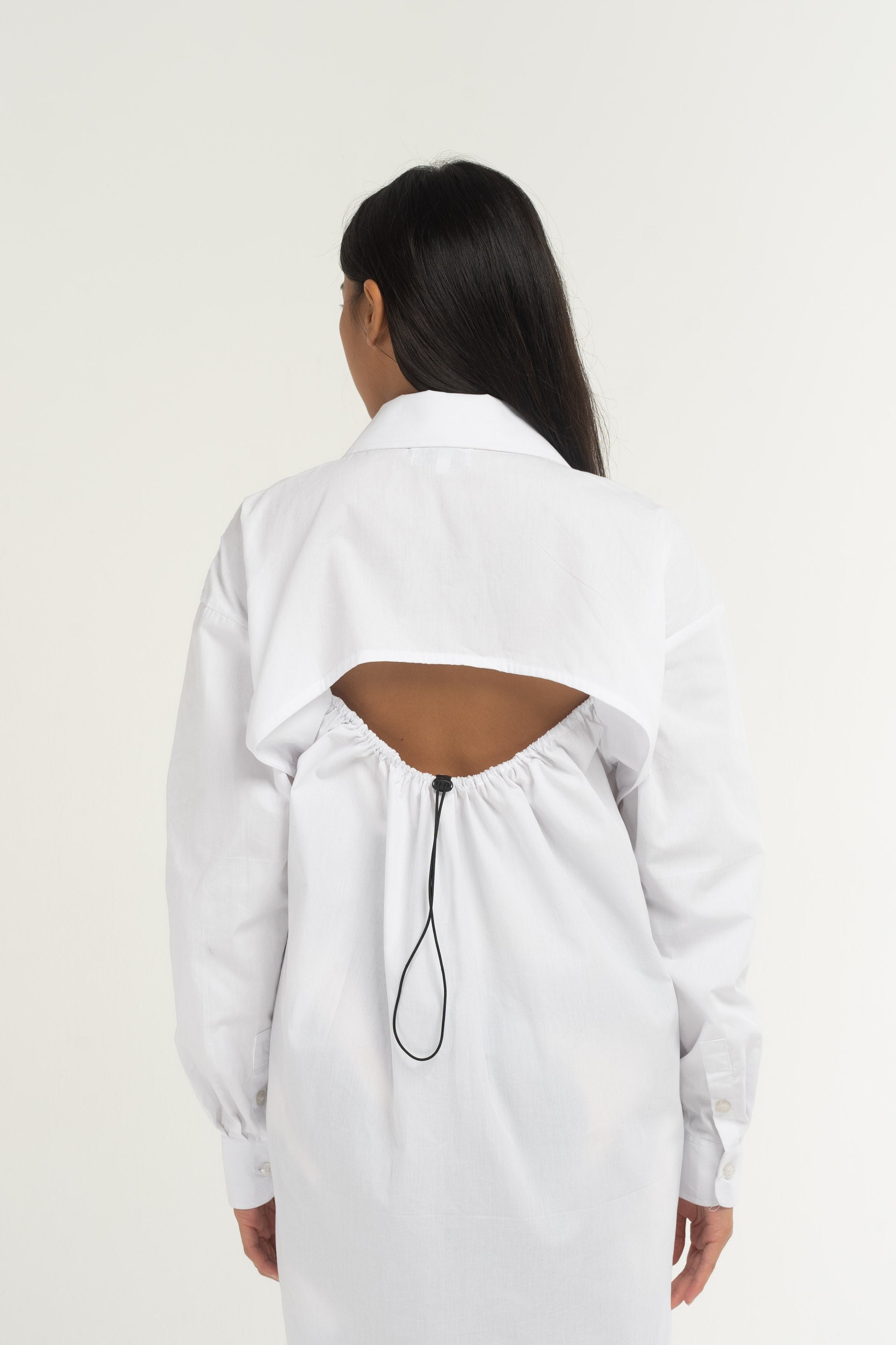 A white Toggle Shirt Dress forms a long silhouette with a curved hem, featuring the Sweet Rose logo hand-embroidered on the front, long-sleeves with a button-up front, a toggle fastening at the back, a structured collar and cuffs.